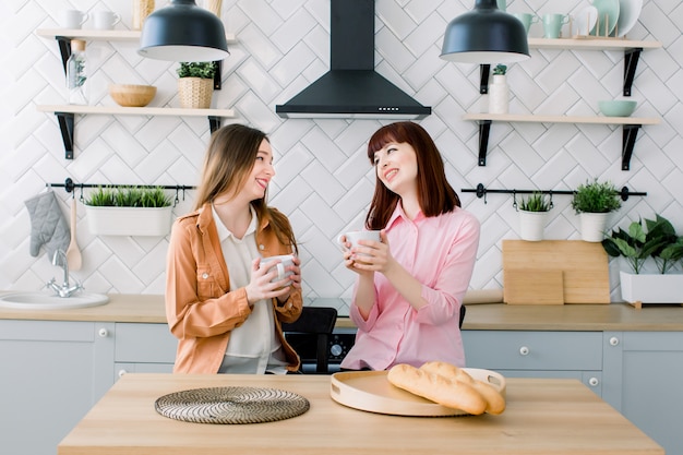 Two brunette girls friend drinking coffee or green tea talking. Couple of woman eating breakfast together on kitchen interior background