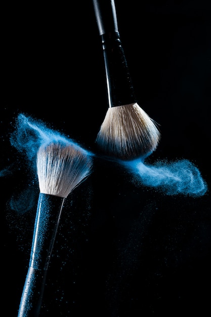 Две кисти для макияжа с голубой макияж тени в движении на черном фоне.