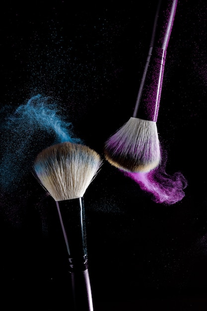 Две кисти для макияжа с синей и розовой макияжа тени в движении на черном фоне.