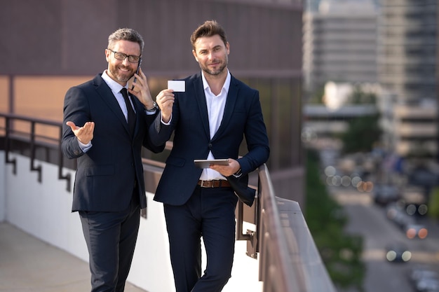 Two business men in suit hold credit card and laptop tablet outdoor pay the bill man with plastic cr