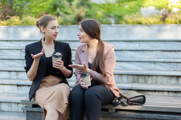 Две подруги-деловые женщины сидят на скамейке, пьют кофе и разговаривают на открытом воздухе