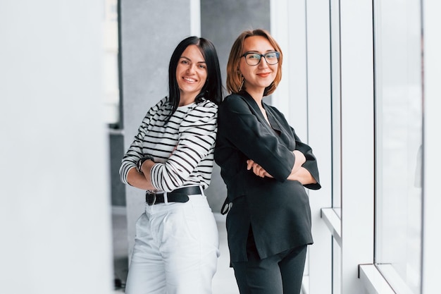 Two business women standing together indoors in the office near big window