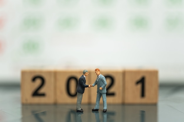 Two businessman miniature figure people make a hand shake with wooden number 2021  block and calendar as background.