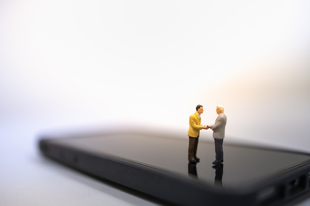 Two businessman miniature people figures standing, talking and hand shake on smart mobile phone.