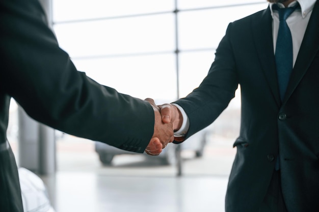 Two businessmen are standing in the car showroom and making a deal handshake