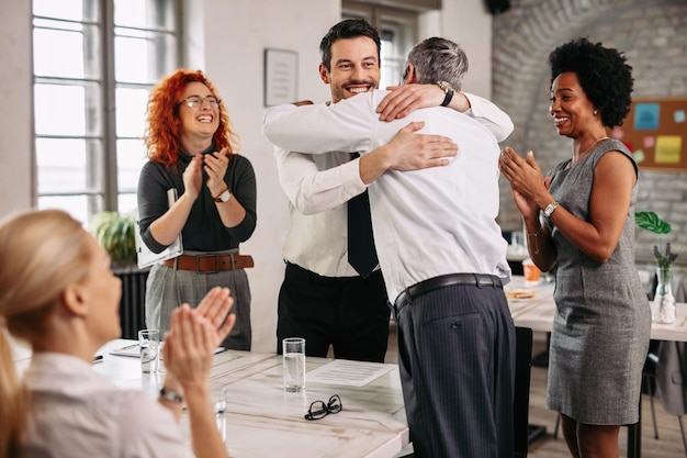 Two businessmen congratulating each other on successful job and embracing during a meeting in the office Other colleagues are applauding them