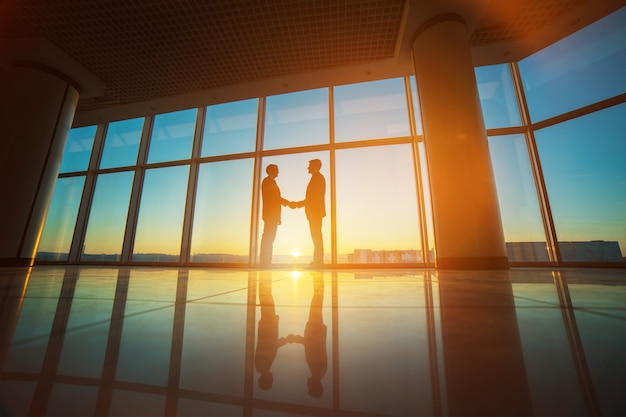 The two businessmen handshake in the office on the bright sun background