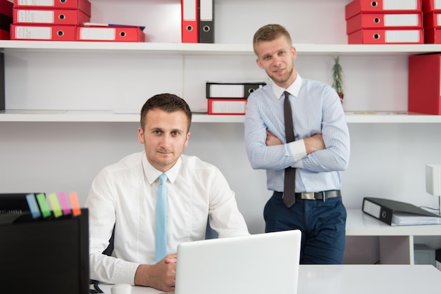 Two Businessmen In An Office Smiling At The Camera While Working Together Behind A Laptop Computer