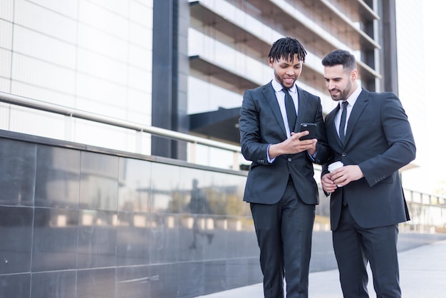 Two businessmen wearing suits walking and using phone smiling and talking outdoor in a business area