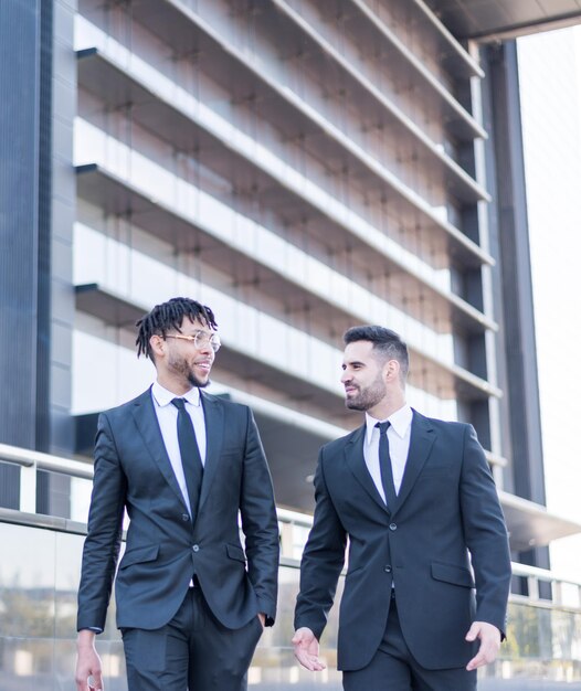 two businessmen wearinng suits walking and using phone smiling and talking outdoor in a business area