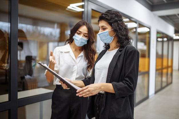 Two businesswomen discuss working matters at the office wearing medical sterile masks. Working during coronavirus pandemic quarantine.