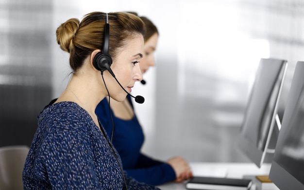 Two busineswomen have conversations with the clients by headsets, while sitting at the desk in a modern office. Diverse people group in a call center. Telemarketing and customer service