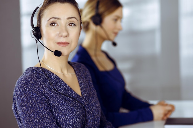 Two busineswomen have conversations with the clients by headsets, while sitting at the desk in a modern office. Diverse people group in a call center. Telemarketing and customer service.