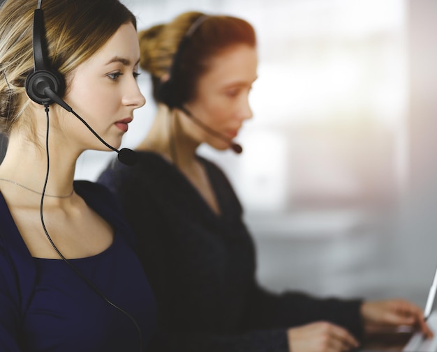 Two busineswomen have conversations with the clients by headsets, while sitting at the desk in a sunny modern office. Diverse people group in a call center. Telemarketing and customer service