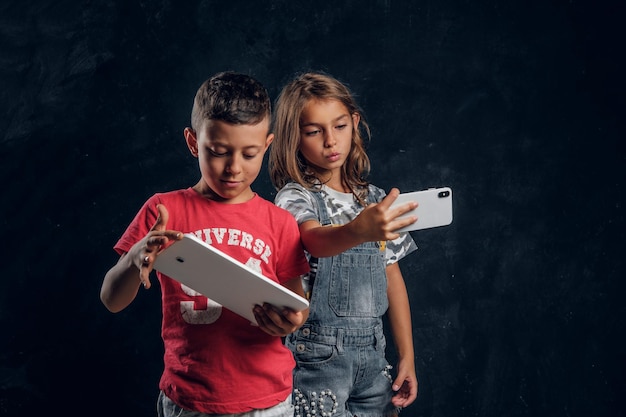 Two busy kids - boy is playing games on tablet while his sister is making a selfie by mobile phone.