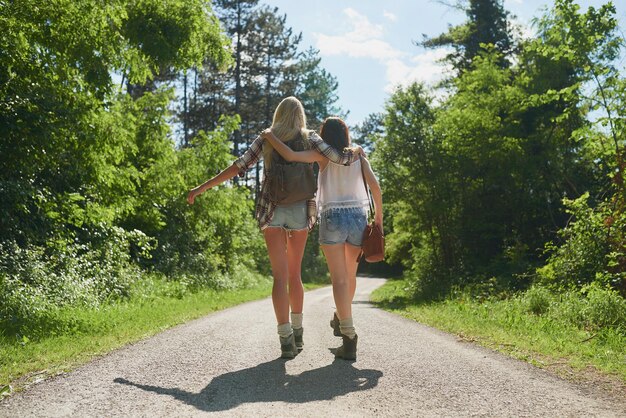 Two carefree women walking and hugging each other during a hike Carefree friends on holiday hugging during a hike Two friends walking down a road in a forest during a hike hugging