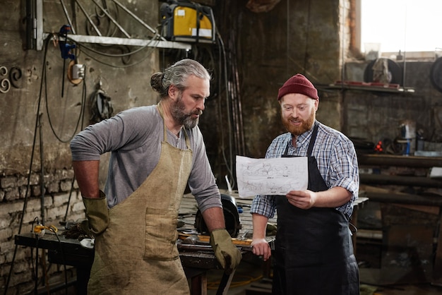 Two carpenters examining blueprint of new product together in team during their work in workshop