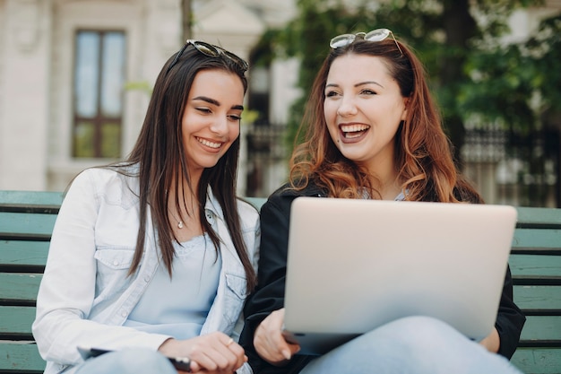 Due bella femmina caucasica che si siede su una panchina che ride guardando un laptop contro un edificio.