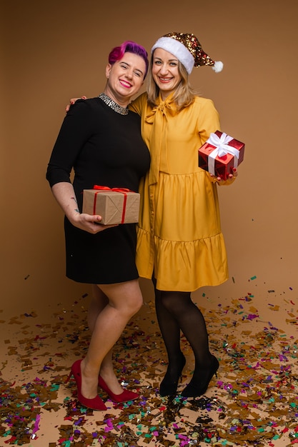 Photo two caucasian beautiful women black and yellow dresses celebrates new year together with presents, picture isolated on brown wall