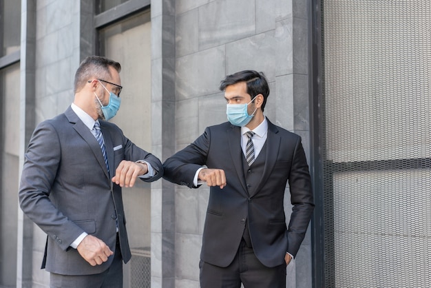 두 백인 사업가가 거리에서 코로나 바이러스 COVID-19 전염병 동안 팔꿈치를 부딪쳐 의료 마스크 인사말을 착용합니다.