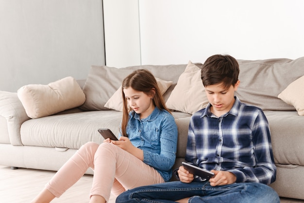 two caucasian children girl and boy sitting on floor near sofa at home, and both using smartphone