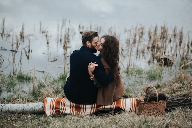 Two caucasian lovers sitting on blanket by the lake. Young couple is hugging on autumn day outdoors. A bearded man and curly woman in love.