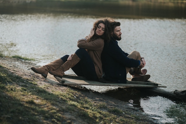 Two caucasian lovers sitting on the pier by the lake. A bearded man and curly woman in love. Valentine's Day.