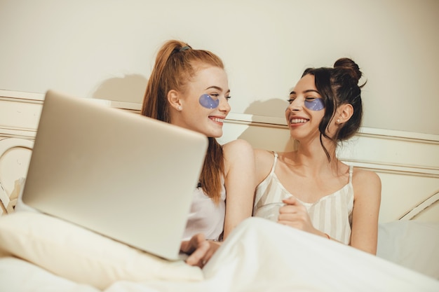컴퓨터와 함께 침대에 누워있는 동안 노화 방지 마스크를 쓰고 두 백인 자매