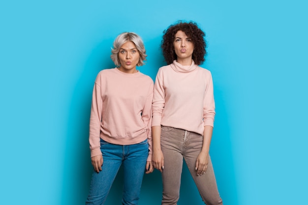 Two caucasian sisters with curly hair wearing the same sweater are posing and gesturing with lips a kiss on a blue wall