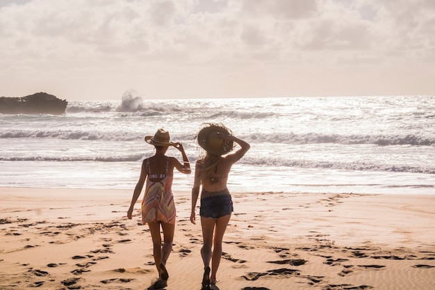 Foto due donne caucasiche con cappello turistico viste da dietro a piedi al mare con le onde