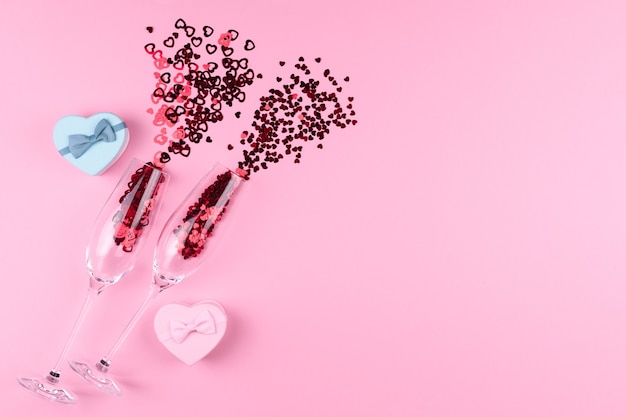 Two champagne glasses filled with heart shaped sequins with a bottle of champagne and a Valentine's Day gift on a pink background.