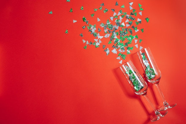 Two champagne glasses with splashing confetti in the form of Christmas trees on a red background. New Year and Christmas concept. Copy space