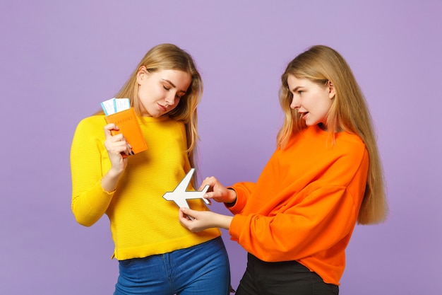 Two charming blonde twins sisters girls in colorful clothes hold passport, boarding pass ticket, paper airplane isolated on violet blue wall. People family lifestyle concept. 