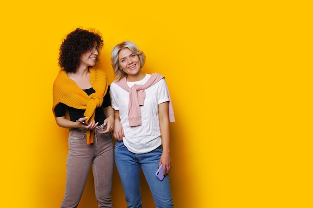 Two charming curly haired sisters are posing on a yellow  wall smiling at front near free space