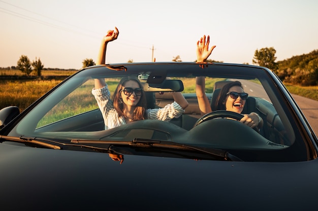 Two charming dark-haired young women in sunglasses are sitting in a black cabriolet on a sunny day. .