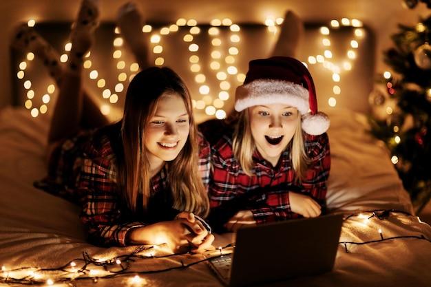 Two charming sisters lie on the bed with a laptop on christmas or new years eve