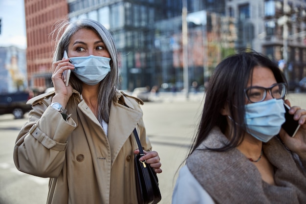 Photo two charming women in medical masks talking on cellphone outdoors