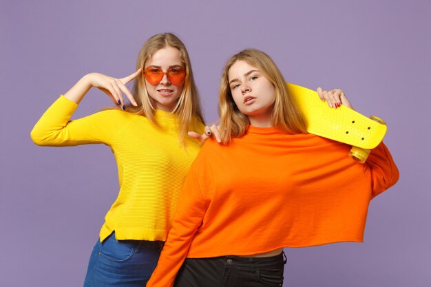 Two charming young blonde twins sisters girls in vivid clothes, heart eyeglasses hold yellow skateboard isolated on pastel violet blue wall. People family lifestyle concept. 