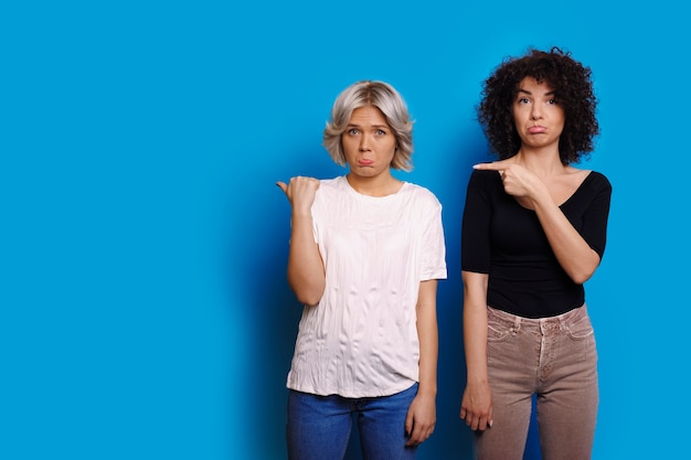 Photo two charming young woman dressed in white and black pointing at the left with fingers and looking questioned at the camera isolated on blue wall.