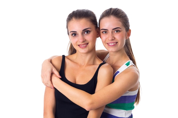 Two charming young women in striped and black shirts hugging on white background in studio