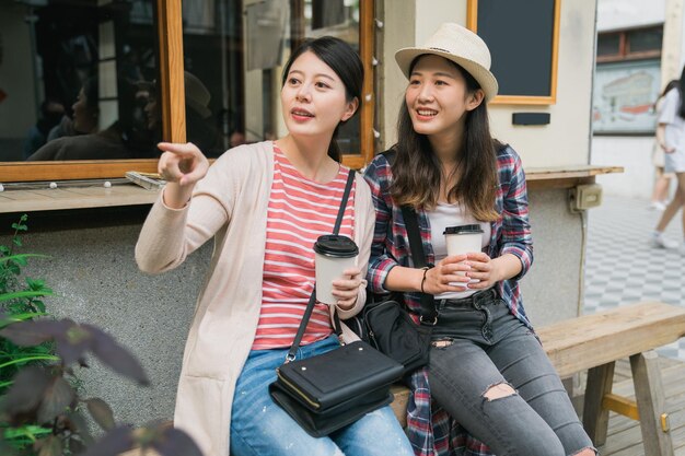 Two cheerful asian korean women traveler resting at summer cafe\
street outdoors while shopping in little village weekend market.\
girl friends discussing next tourist attraction with finger point\
way