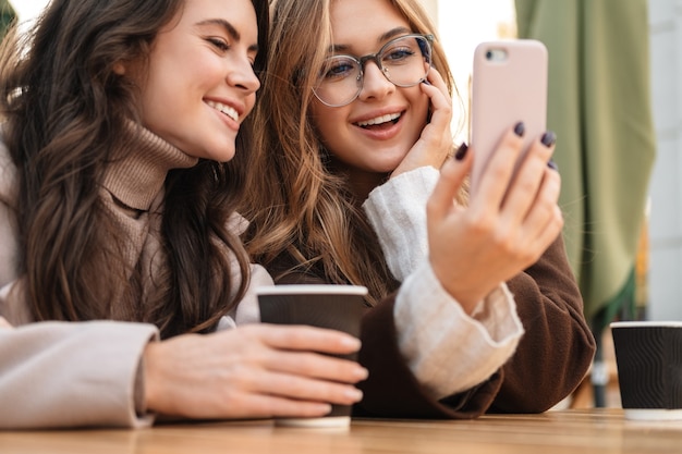 Two cheerful attractive women friends sitting at the cafe outdoors, looking at mobile phone while drinking coffee