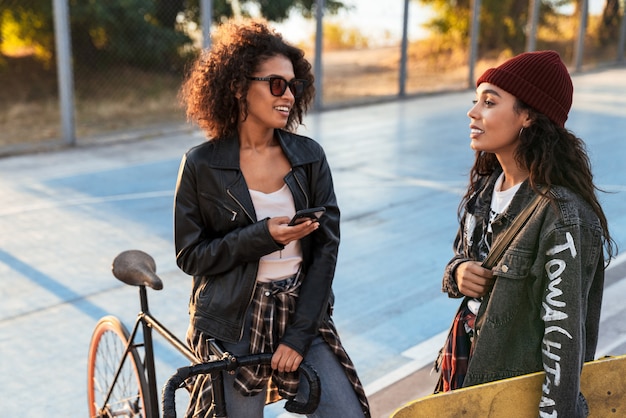 Two cheerful attractive young african girls standing at the sports ground with bicycle and skateboard, using mobile phone