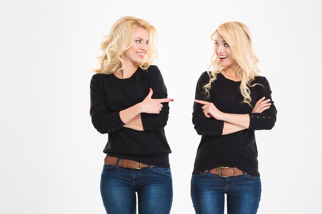 Two cheerful attrative sisters twins pointing and looking at each other over white background