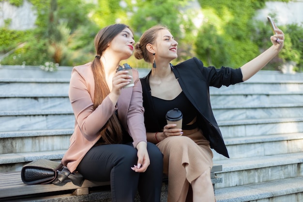 Two cheerful business women take a selfie on a smartphone while sitting on a bench in the park