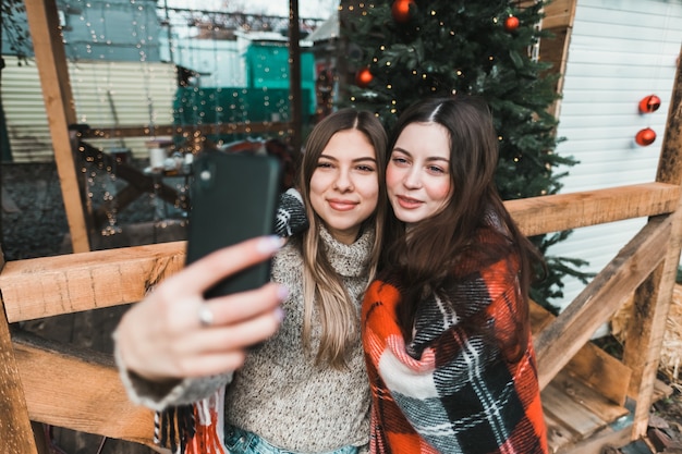 Two cheerful caucasian women friends having fun and making selfie on the backyard with Christmas decorations.