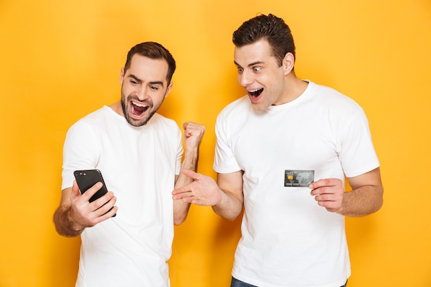 Due amici uomini allegri eccitati che indossano magliette bianche in piedi isolate su un muro giallo, guardando il telefono cellulare, celebrando il successo