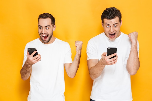 Two cheerful excited men friends wearing blank t-shirts standing isolated over yellow wall, using mobile phones, celebrating success