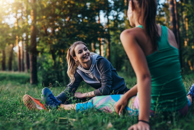 Two cheerful female athletes warming up before training doing stretching exercise for legs sitting on grass