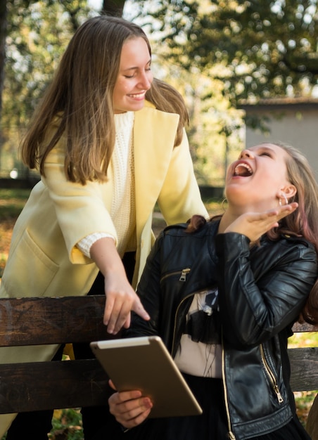 Two cheerful female best friends in casual wear using tablet in autumn sunny park Happy teen girls outdoors